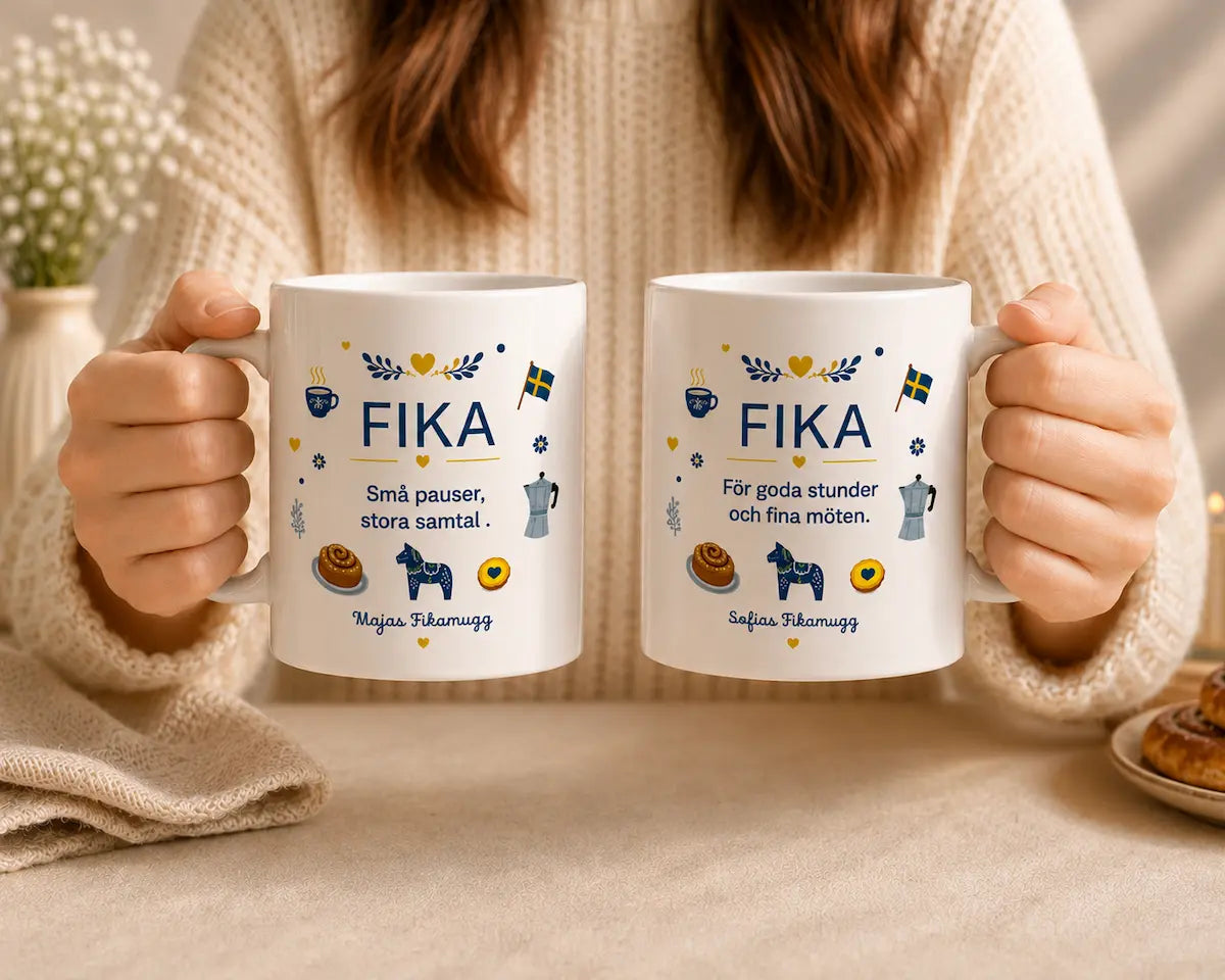 Two mugs with 'FIKA' text and illustrations held by a person wearing a beige sweater.